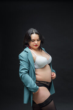 A beautiful girl in a green jacket and black stockings stands on a black background. She is wearing a white bra and red lipstick.