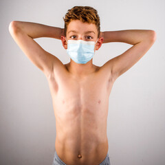 Young Caucasian boy standing shirtless in a medical face mask with arms up, hands behind his head