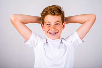 Head and shoulders portrait of a preteen boy with freckles and auburn hair, smiling with braces, while posing with his hands behind his head - obrazy, fototapety, plakaty