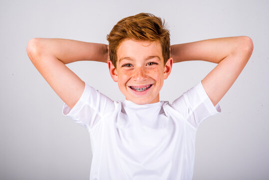 Head and shoulders portrait of a preteen boy with freckles and auburn hair, smiling with braces, while posing with his hands behind his head