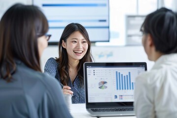 Three Japanese office workers are smiling and discussing work in the meeting room, with a laptop displaying financial data on the screen