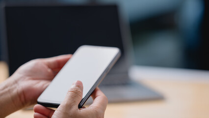 A person is holding a cell phone in their hand, with a laptop in the background. Concept of productivity and focus, as the person is likely working on their phone or laptop