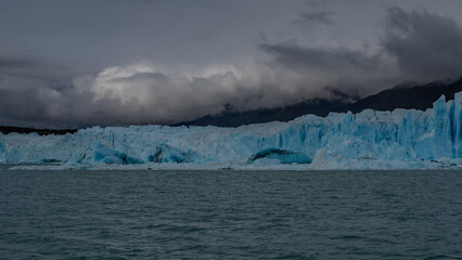 A wall of blue ice stretches across the glacial lake.  Broken icebergs, melted ice floes float in...