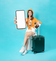 Happy Asian woman traveler sitting on the chair and using smartphone with holding passport and plane tickets with suitcase isolated on blue background.