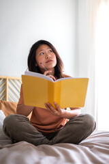 Teen asian girl sitting on bed thinking, using diary and pen to write down thoughts and ideas. Vertical.