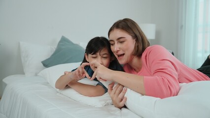 Happy asian daughter and smart mom lie on bed while looking at camera. Energetic family spend time and prepare to sleep together. Cute girl and mom smile to camera show love and affection. Pedagogy. - obrazy, fototapety, plakaty