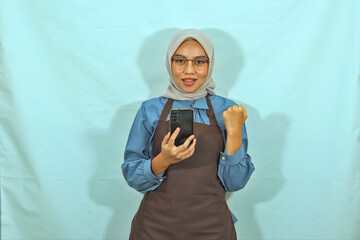 Asian Muslim woman wearing hijab, glasses and brown apron holding mobile phone while making a winner gesture, smile showing her teeth isolated on a white background. housewife muslim lifestyle concept