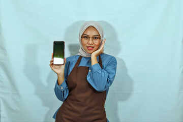 young Asian Muslim woman wearing hijab, glasses and a brown apron showing a blank screen mobile phone isolated on a white background. housewife muslim lifestyle concept
