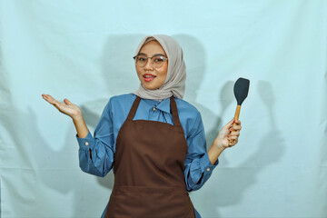 Asian Muslim woman wearing glasses and a brown apron holding a spatula and spreading her hands...
