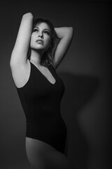 Black and white portrait of a beautiful caucasian model wearing a black bodysuit. Her arms are raised into a pose. This is photographed in the studio with a dark background.