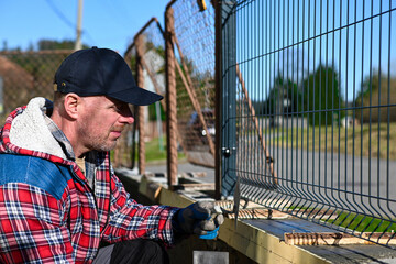 A man in overalls and gloves is repairing the fence in front of the family house. Close-up view and...