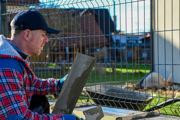 A man in overalls and gloves is repairing the fence in front of the family house. He uses a plane,...