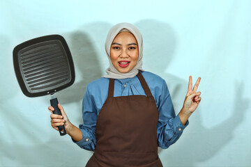 beautiful Asian woman in brown apron and hijab holding pan and showing peace sign with fingers....
