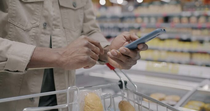 Close-up of male hand using smartphone and checking products in shopping trolley while man buying food in supermarket. Modern gadget and retail concept.