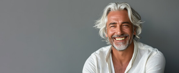 A man with white hair and a white shirt is smiling. He is sitting on a grey surface. Attractive 50 year old smiling man with medium white hair in cream-colored clothes against a gray background. - obrazy, fototapety, plakaty