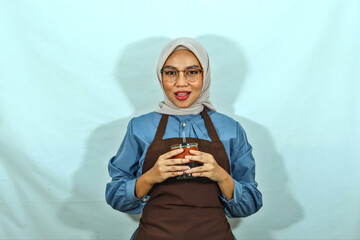 young Asian muslim woman wearing hijab, glasses and brown apron coffee cup with hands. smile showing her teeth isolated over white background. housewife muslim lifestyle concept
