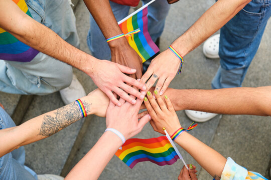 A group of people stacking hands and holding rainbow flags. Scene is one of unity and support for the LGBTQ community.