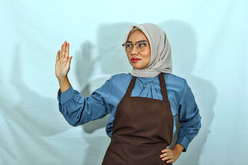 unhappy young Asian Muslim woman in hijab, glasses and brown apron showing stop gesture refusing...