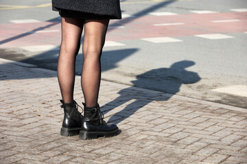 Female legs in black boots with shadow on pavement. Slim girl in pantyhose standing on a street, fashion in spring city