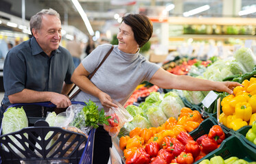 Smiling happy couple is choosing bell peppers and other vegetables in supermarket - obrazy, fototapety, plakaty