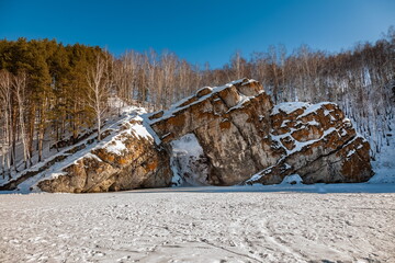 Winter landscape with a large stone on the bank of a frozen river , forest and sky