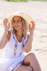 Young beautiful girl with blond hair in a summer dress and hat sits on the sand on the shore, portrait, pose.