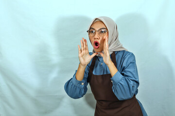 young Asian Muslim woman wearing hijab, glasses and brown apron, screaming shouting good news with hands near mouth isolated on white background. housewife muslim lifestyle concept
