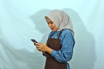 young Asian muslim woman wearing hijab and brown apron typing messages in mobile phone isolated...