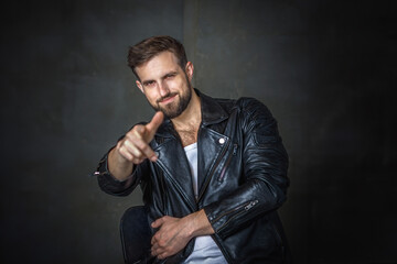 Modern young man with a stylish hairstyle, wearing a cool black leather jacket and sitting on a chair on a dark background
