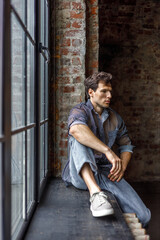 A modern young man with a stylish hairstyle sits on a windowsill near a window on a dark background