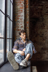 A modern young man with a stylish hairstyle sits on a windowsill near a window on a dark background