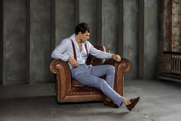 A young businessman is thinking after a meeting, posing in front of the camera in business formal clothes, sitting on an armchair