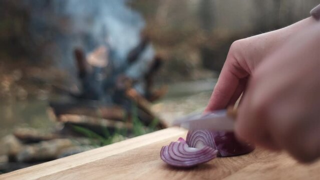 Close up of female hands cutting fresh purple onions on wooden board near warm bonfire and river in evening spring time. Healthy eating. Camping vibes, nature and outdoor lifestyle mood concept