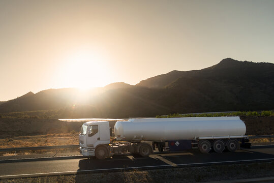 White fuel tanker truck driving on a highway with the sun rising between the mountains, side view.