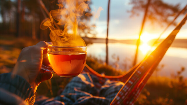 a cup of hot tea with steam in the hands of a man sitting in a hammock against the backdrop of the setting sun --ar 16:9 --style raw Job ID: e71a6734-a126-45dc-bb6e-462b801ce8b3