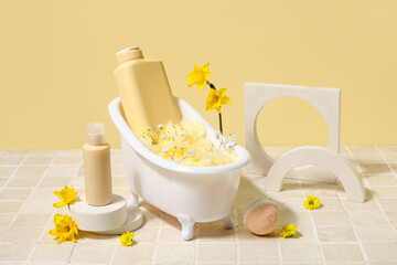 Composition with mini bathtub, bottles of cosmetic products and flowers on tile table near yellow wall