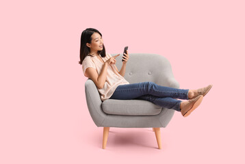 Beautiful Asian woman using mobile phone in grey armchair on pink background - obrazy, fototapety, plakaty