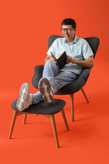 Young man reading book in armchair with footrest on red background
