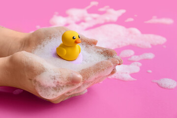Female hands with soap foam and yellow rubber duck toy on pink background
