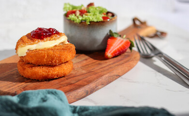 A portion of fresh made fried camembert cheese with cranberry sauce on top and a side of fresh salad on wooden plate