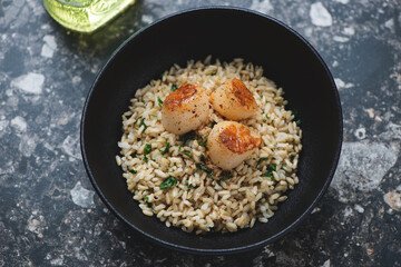 Seared scallops with brown rice, lemon zest and parsley served in a black bowl, horizontal shot on a dark-brown granite background