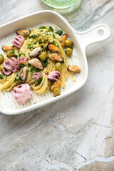 Seafood spaghetti with green olives, parsley and lemon zest in a beige serving tray, vertical shot on a grey granite background with space