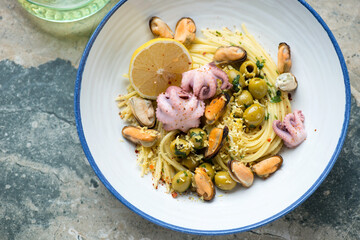 Spaghetti with mussels, baby octopuses and green olives in a blue and white plate, horizontal shot on a grey and beige granite surface, high angle view