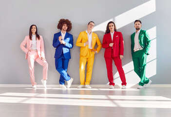 Group of positive young people in colorful business suits standing against a gray wall. They...