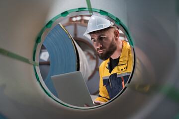 Male engineer worker using laptop computer inspecting quality rolls of galvanized or metal sheet in...
