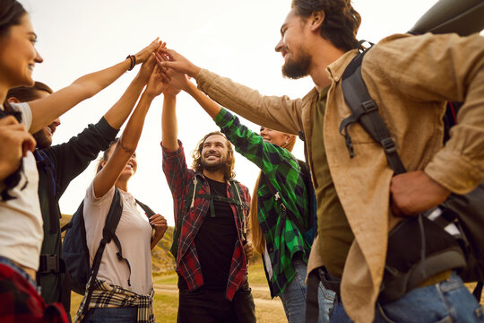 Group of happy tourist friends with backpacks raises hands, a symbolic gesture of triumphant teamwork during vacation. This team showcases the essence of joyous travel in nature and friendship.