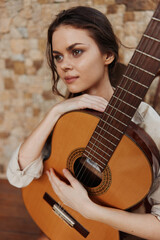 A young woman holding an acoustic guitar in front of a stone wall and looking at the camera