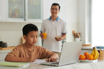 Vietnamese schoolboy watching educational video on laptop and doing homework