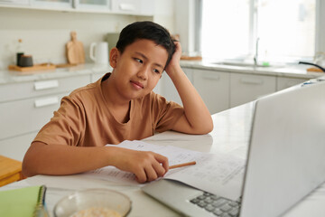 Pensive Vietnamese schoolboy reading article on laptop screen when doing math homework