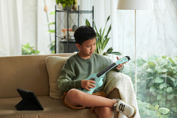Pensive preteen boy trying to play ukulele at home